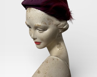 May include: A vintage purple hat with a structured crown and a small brim. The hat is adorned with a cluster of dark red feathers on the side. The hat is displayed on a mannequin head with a distressed, aged finish.