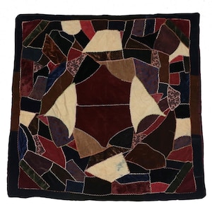 May include: A square patchwork quilt with a black border. The quilt is composed of various fabric shapes in shades of red, brown, black, and cream. The pieces are sewn together with white stitching, creating a geometric pattern.