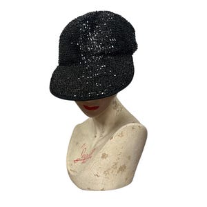 Puede incluir: Una gorra de béisbol negra con lentejuelas y visera curva. La gorra está sobre una cabeza de maniquí blanca.
