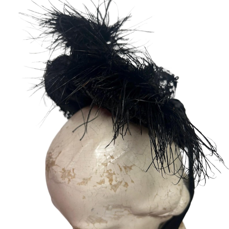 May include: A black fascinator with a cluster of feathers and lace detailing. The headpiece is mounted on a small base, creating a dramatic and elegant look. The feathers add texture and movement to the design.