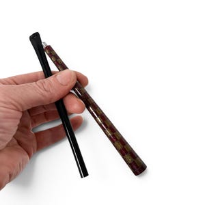 May include: Two slender, cylindrical writing instruments. One pen features a solid, glossy black finish. The second pen displays a decorative, alternating checkered pattern in deep burgundy and shimmering gold tones along its entire length.