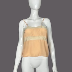 May include: A peach-colored camisole top with thin spaghetti straps. The top features a band of cream-colored lace across the bust. The garment is displayed on a white mannequin against a gray background. The top is made of a lightweight, flowing fabric.