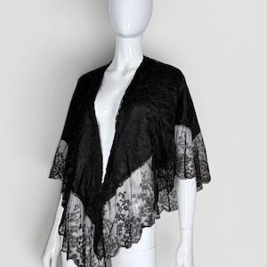 May include: A black lace shawl featuring an intricate floral pattern throughout the fabric. The garment has an open front design with a scalloped edge along the hemline and sleeves, providing a lightweight, semi-sheer layered appearance.