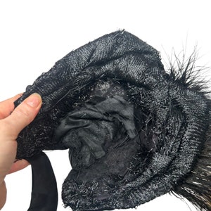 May include: A black hat with a textured, woven design and a black satin ribbon. The hat is adorned with black feathers. The interior of the hat is visible, revealing a black lining.