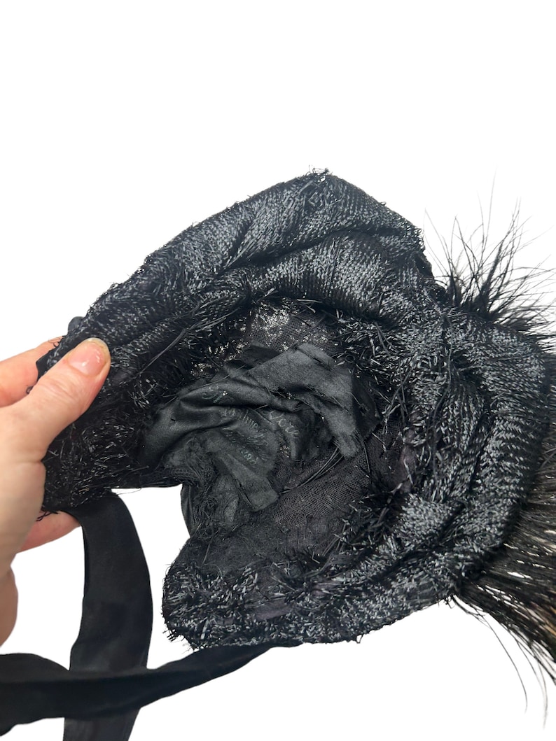 May include: A black hat with a textured, woven design and a black satin ribbon. The hat is adorned with black feathers. The interior of the hat is visible, revealing a black lining.