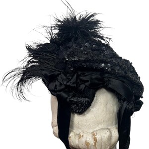 May include: A black hat with sequins, feathers, and a satin bow. The hat has a rounded crown and a wide brim. Long black ribbons hang from the hat. The hat is likely vintage or antique.