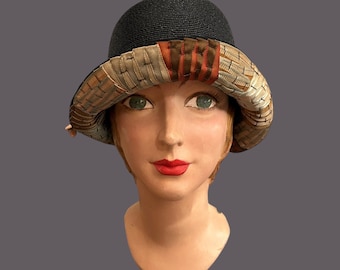 May include: A black cloche hat with a woven straw crown and a decorative brim. The brim features a woven pattern in shades of beige, brown, and orange. The hat is displayed on a mannequin head.