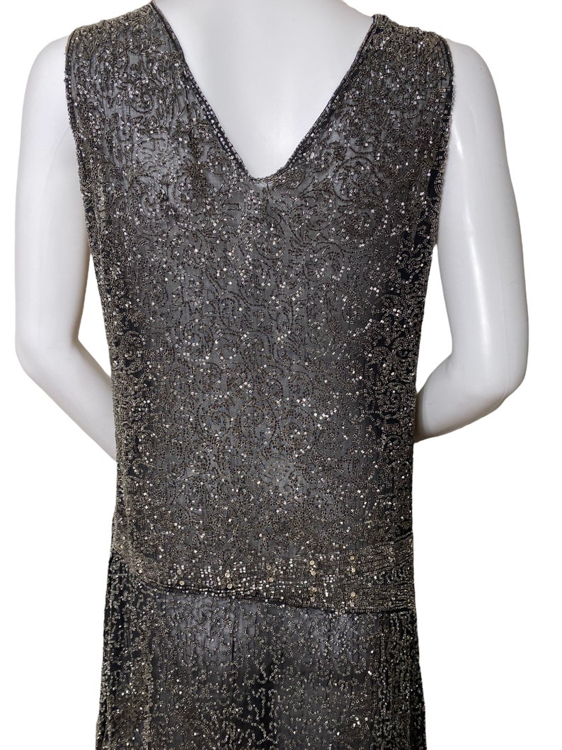 May include: A sleeveless, beaded dress in a dark gray color. The dress has a V-neckline and a beaded pattern throughout. The dress is fitted through the bodice and flows out at the hem.