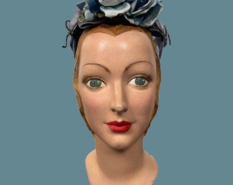 May include: A vintage style mannequin head with a blue and white flower hat. The mannequin has blonde hair and green eyes. The hat is made of fabric and has a blue ribbon.