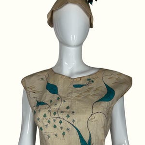May include: A beige map-printed top and matching hat on a mannequin. The sleeveless top features a teal river design and tree illustrations. The hat is cone-shaped with a dark green accent.