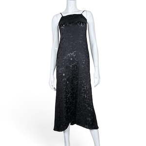 May include: A black midi-length slip dress featuring a tonal floral pattern throughout. The garment has a straight neckline, thin spaghetti straps, and a fluid, straight-cut silhouette that falls below the knees, displayed on a white mannequin.