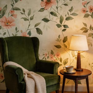 May include: A cozy interior scene featuring a dark green velvet armchair with a cream-colored throw blanket. A small wooden side table holds a lamp with a beige lampshade and a teacup. The background is a floral wallpaper with pink flowers and green leaves.