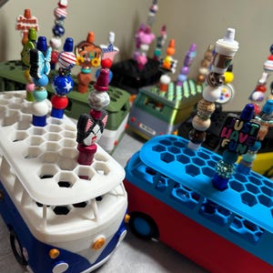 May include: A collection of colorful van-shaped pen holders with honeycomb tops, each holding multiple decorative beaded pens. The pens feature various charms, including a cat face, a coffee cup, and the text "JUST HAVE FUN" in block letters.