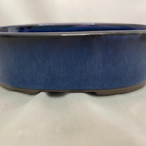 May include: An oval-shaped ceramic bonsai pot featuring a deep blue glazed finish with subtle variations in tone. The rim is smooth and rounded, while the base is unglazed, revealing a natural brown clay texture along the bottom edge.