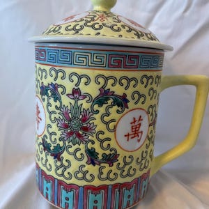 May include: Yellow ceramic mug with a matching lid. The mug features a floral design with red, pink, and blue accents. The handle and lid knob are yellow. The mug has Chinese characters on the side.