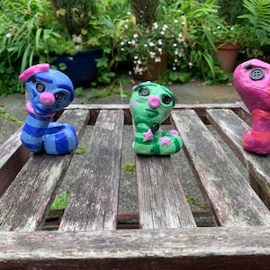 May include: Three handmade clay figurines shaped like coiled creatures. The left one is blue with stripes, the centre one is green with stars, and the right one is pink. Each figure features black button eyes, a pink nose, and painted facial details.