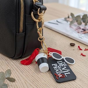 Può includere: Una borsa a tracolla nera con catena e cerniera dorate. Alla catena sono attaccati ciondoli: una scarpa con tacco rosso, una tazza di caffè, occhiali da sole bianchi e un'etichetta nera con "The Devil Wears Prada".