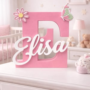 Può includere: Una lettera "E" rosa in legno con il nome "Elisa" in corsivo bianco. La lettera è decorata con una margherita e una farfalla. L'arredamento è posto su un comò bianco, che suggerisce una cameretta.