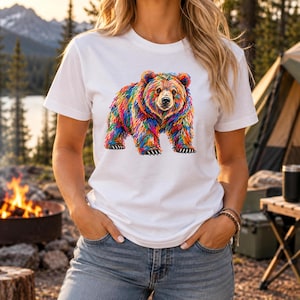 May include: A white short-sleeved t-shirt featuring a graphic print of a bear rendered in vibrant, multi-colored brushstrokes. The bear is depicted in a standing position, showcasing a spectrum of colors including red, blue, yellow, and green.
