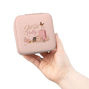 May include: A light pink travel jewelry box with the words "Jet Set Pretty" in gold script. The box features a graphic of a pink suitcase, purse, sunglasses, and flowers. A small butterfly is also included in the design.