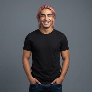 Puede incluir: Una camiseta negra de cuello redondo con un pequeño logotipo en el pecho. La camiseta está hecha de una tela oscura y suave. La persona lleva vaqueros azules y tiene el pelo rosa. El fondo es gris liso.