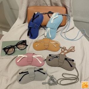 May include: A selection of spectacle cases in shades of blue, light blue, pink, grey, and tan. Each case features a strap and a snap closure. A pair of black-framed glasses and a small brown bag are also present.