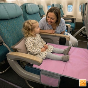 Peut inclure: Un lit de voyage rose pour enfants installé sur un siège d'avion. Une jeune enfant, vêtue d'une robe à fleurs et de leggings gris, est assise sur le lit. Une femme est assise à côté, souriant à l'enfant.