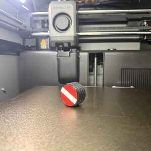 May include: A small, cylindrical 3D-printed object with a black body and a red and white stripe. The object is positioned on a flat, textured surface inside a 3D printer.