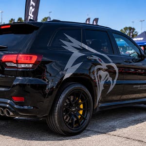 May include: A black Jeep Grand Cherokee Trackhawk SUV featuring a large grey Hellcat logo decal on the rear side panel, black multi-spoke wheels with yellow brake calipers, dual exhaust pipes, and red taillights under a clear blue sky.