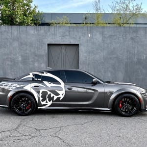 May include: A dark gray sedan featuring a large white graphic of a stylized cat head on the rear side panel. The vehicle has black multi-spoke wheels, red brake calipers, tinted windows, and a rear spoiler. The car is parked on asphalt.