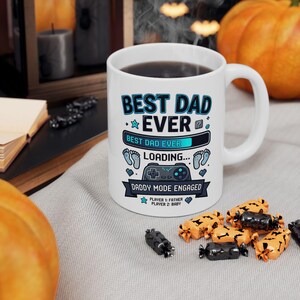 Peut inclure: Tasse en céramique blanche avec l'inscription "BEST DAD EVER" en bleu et noir. La tasse présente un graphique de barre de chargement, des empreintes de pas et une manette de jeu. Le texte "DADDY MODE ENGAGED" est également présent. La tasse est remplie d'un liquide foncé.
