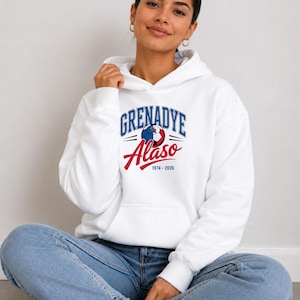Peut inclure: Un sweat-shirt à capuche blanc avec l'inscription "GRENADYE Alaso 1974-2026" en bleu et rouge. Le design comprend un graphisme de ballon de football. La personne porte un jean bleu.