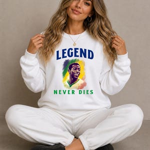 Peut inclure: Sweat-shirt blanc avec l'inscription "LEGEND NEVER DIES" en bleu et vert. Le sweat-shirt présente un portrait d'un joueur de football. La personne sur l'image porte le sweat-shirt.