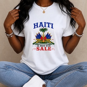 Peut inclure: T-shirt blanc avec le mot "HAITI" en bleu au-dessus d'un emblème coloré et le texte "IS NOT FOR SALE" en rouge. Le t-shirt est porté avec un jean bleu et des baskets blanches.