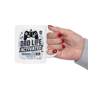 Peut inclure: Tasse en céramique blanche avec un motif bleu et noir indiquant "DAD LIFE ACTIVATED". Le design comprend une manette de jeu, un biberon et le texte "BABY MODE: ON". La tasse est tenue par une personne.