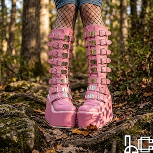 May include: Pink platform boots with multiple silver and gold buckles. The boots have a chunky platform sole and extend to the knees. The boots are made of a smooth material and are worn with fishnet stockings.