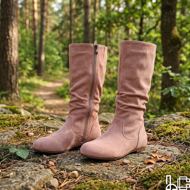 May include: A pair of dusty rose-coloured boots with a side zip. The boots are made of a soft, suede-like material and have a flat sole. The boots are displayed outdoors on a rocky surface with a forest background.