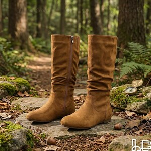 May include: A pair of brown suede knee-high boots with a side zip. The boots have a round toe and a low heel. The boots are displayed on a stone surface in a natural outdoor setting.