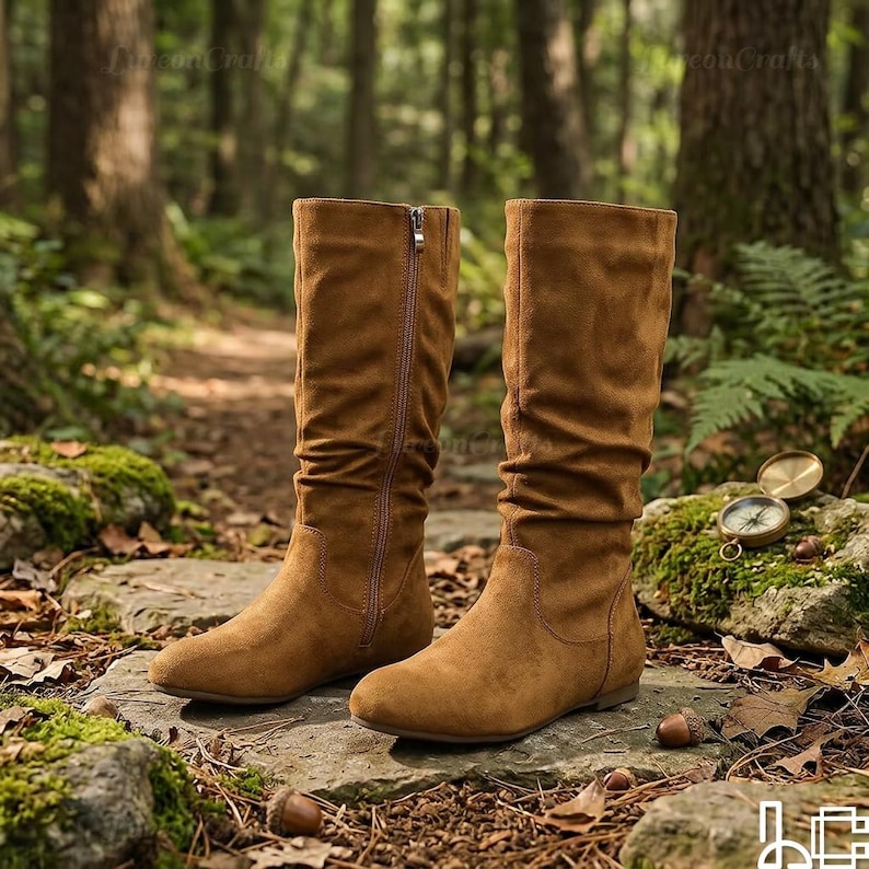 May include: A pair of brown suede knee-high boots with a side zip. The boots have a round toe and a low heel. The boots are displayed on a stone surface in a natural outdoor setting.