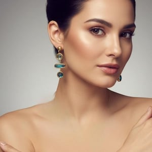 May include: Elegant gold-toned earrings with three teal-colored, geometric-shaped stones. The earrings are dangling and feature a round stud at the top. The woman in the image is wearing the earrings.