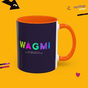 Puede incluir: Una taza de cerámica de color morado oscuro con interior y asa de color naranja brillante. El frente presenta el texto WAGMI en letras multicolores, seguido del texto blanco más pequeño: rug, rugged, fucked over, still here, still holding, not leaving.