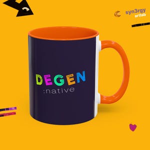 May include: A dark purple ceramic mug with a bright orange interior and handle. The front features the word DEGEN in multicolored, textured block letters above the word :native in a simple white font. The background is a solid yellow color.