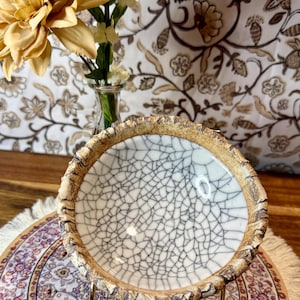 Puede incluir: Un cuenco de cerámica con interior de esmalte craquelado blanco y gris y un borde marrón texturizado. El cuenco se apoya en un soporte de metal dorado, con un fondo de estampado floral y un ramo de flores amarillas en un jarrón de cristal.
