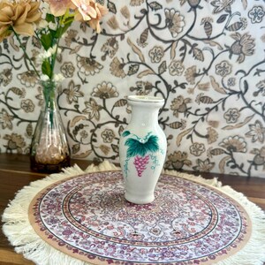 May include: A small, white ceramic vase with a grape design, set on a round, fringed mat with a floral pattern. The vase is decorated with green leaves and a cluster of pink grapes. A glass vase with flowers is in the background.