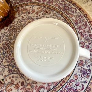 Może przedstawiać: Biały ceramiczny kubek z uchwytem, z napisem "MACKENZIE-CHILDS" i instrukcjami pielęgnacji. Widoczna jest podstawa kubka z tekstem wskazującym, że nadaje się do zmywarki i wyprodukowano w Chinach. Kubek stoi na wzorzystym dywanie.