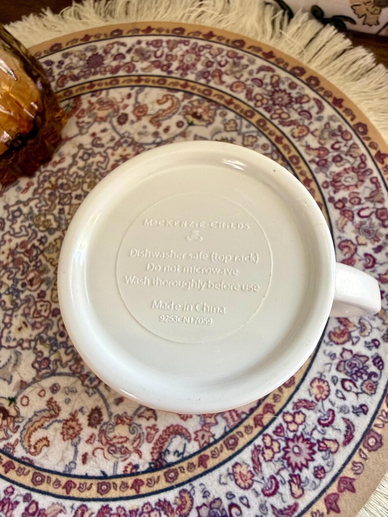 Może przedstawiać: Biały ceramiczny kubek z uchwytem, z napisem "MACKENZIE-CHILDS" i instrukcjami pielęgnacji. Widoczna jest podstawa kubka z tekstem wskazującym, że nadaje się do zmywarki i wyprodukowano w Chinach. Kubek stoi na wzorzystym dywanie.