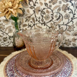 May include: A pale pink pressed glass creamer pitcher featuring a delicate floral pattern and vertical fluting, set upon a matching circular glass plate. The pitcher has a small pouring spout and a curved handle, showcasing a translucent finish.
