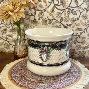 Peut inclure: Un cache-pot en céramique blanche orné d'un motif à pois noirs et blancs avec une bordure décorative de roses roses et de feuilles vertes. Le récipient cylindrique présente un bord évasé et une base arrondie avec un motif floral.