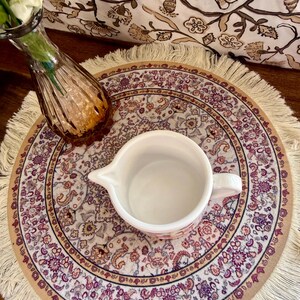 Może przedstawiać: Biała ceramiczna śmietanka z uchwytem stoi na okrągłej, frędzelkowanej podkładce z kwiatowym wzorem w odcieniach fioletu, beżu i brązu. Widoczny jest r&oacute;wnież mały, bursztynowy szklany wazon.