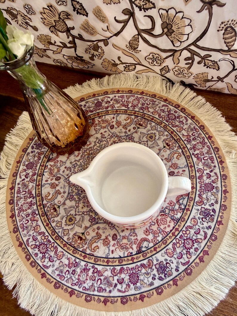 Może przedstawiać: Biała ceramiczna śmietanka z uchwytem stoi na okrągłej, frędzelkowanej podkładce z kwiatowym wzorem w odcieniach fioletu, beżu i brązu. Widoczny jest r&oacute;wnież mały, bursztynowy szklany wazon.
